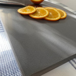 May include: A gray, textured cutting board with a thick edge, featuring a whole orange and several orange slices on top.