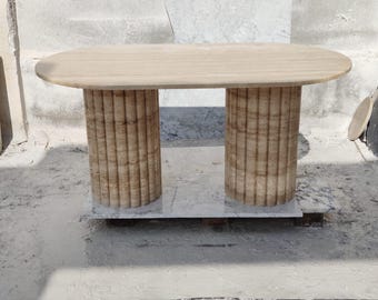 May include: A light brown oval-shaped travertine dining table with two fluted columns for legs. The table is on a white marble base.