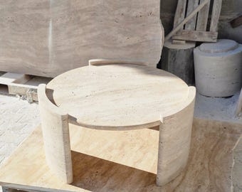 May include: A round, light beige travertine coffee table with a unique, curved design. The table has a smooth, polished surface and a sturdy base.