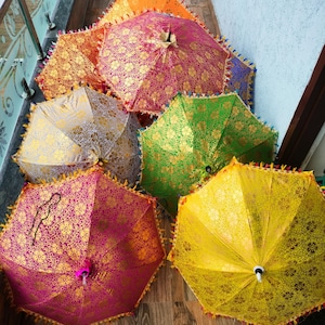 May include: A collection of colorful, open parasols with intricate gold floral patterns. The parasols are in shades of pink, orange, yellow, green, and silver, with decorative fringe around the edges. The parasols are arranged on a wooden surface.