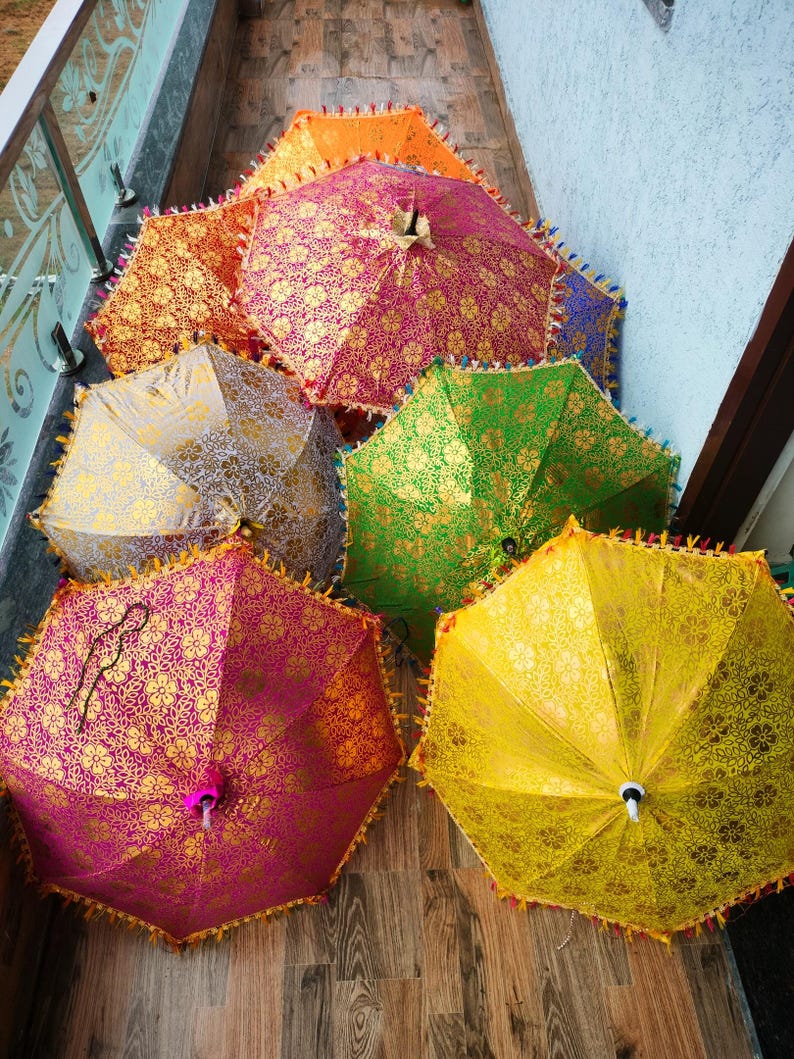 May include: A collection of colorful, open parasols with intricate gold floral patterns. The parasols are in shades of pink, orange, yellow, green, and silver, with decorative fringe around the edges. The parasols are arranged on a wooden surface.