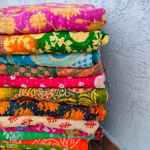 Op de afbeelding: Een stapel kleurrijke, bedrukte quilts. De quilts hebben een verscheidenheid aan kleuren, waaronder roze, groen, oranje en blauw. Elke quilt heeft een uniek ontwerp, met bloemen- en geometrische patronen. De quilts zijn opgevouwen en netjes opgestapeld.