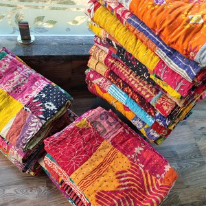 May include: A stack of colorful, patchwork quilts in various shades of red, orange, yellow, pink, and blue. The quilts are folded and stacked, showcasing the intricate stitching and patterns. The quilts appear to be made of soft, textured fabric.
