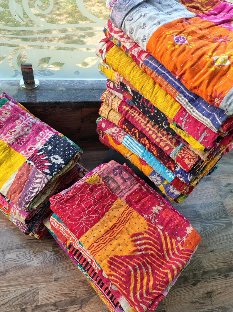 May include: A stack of colorful, patchwork quilts in various shades of red, orange, yellow, pink, and blue. The quilts are folded and stacked, showcasing the intricate stitching and patterns. The quilts appear to be made of soft, textured fabric.
