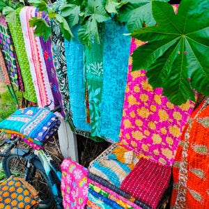 Op de afbeelding: Een display van kleurrijke, bedrukte textiel, waaronder quilts en dekens, hangend aan een houten hek en gestapeld op een fiets. De textiel heeft verschillende kleuren en patronen, waaronder bloemen- en geometrische patronen.