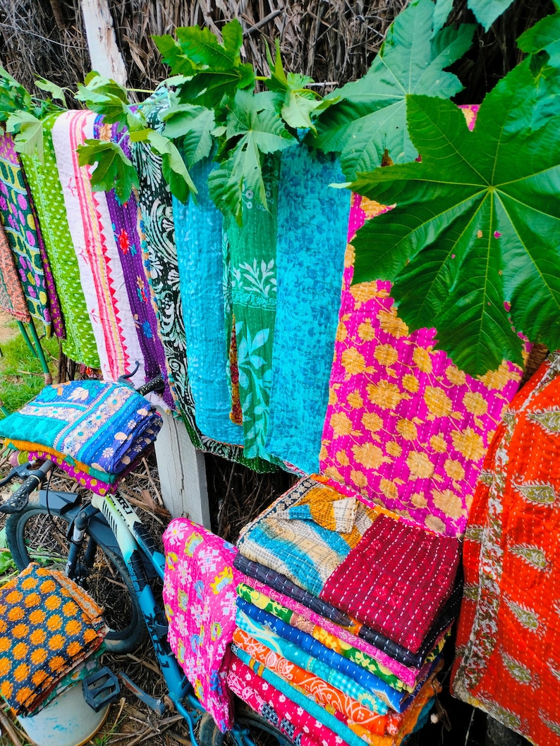 Op de afbeelding: Een display van kleurrijke, bedrukte textiel, waaronder quilts en dekens, hangend aan een houten hek en gestapeld op een fiets. De textiel heeft verschillende kleuren en patronen, waaronder bloemen- en geometrische patronen.