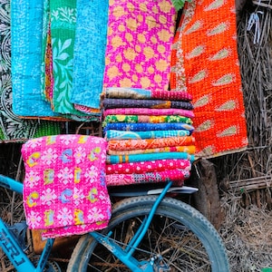 Op de afbeelding: Een display van kleurrijke, bedrukte textiel, waaronder quilts en plaids, zijn opgestapeld en gedrapeerd. Een blauwe fiets staat op de voorgrond, met een roze textiel bevestigd op de achterdrager. De textiel heeft bloemen- en geometrische patronen.