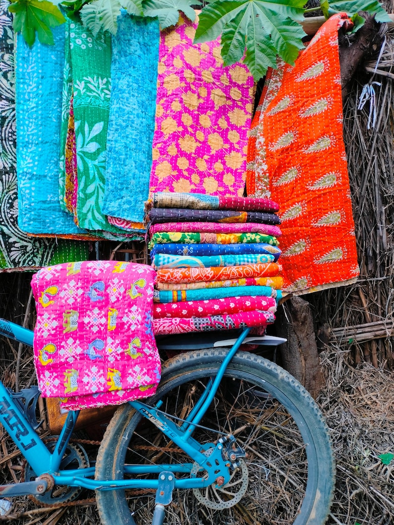 Op de afbeelding: Een display van kleurrijke, bedrukte textiel, waaronder quilts en plaids, zijn opgestapeld en gedrapeerd. Een blauwe fiets staat op de voorgrond, met een roze textiel bevestigd op de achterdrager. De textiel heeft bloemen- en geometrische patronen.