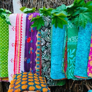 Op de afbeelding: Een verzameling kleurrijke, bedrukte textiel, waaronder quilts en dekens, wordt buiten tentoongesteld. De textielstukken hebben verschillende ontwerpen, waaronder bloempatronen, geometrische vormen en ingewikkelde stiksels. Het kleurenpalet omvat levendige tinten zoals roze, groen en blauw.