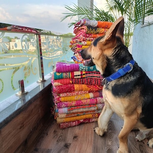 May include: A stack of colorful, patterned quilts in shades of pink, orange, yellow, and purple. A German Shepherd dog with a blue collar sits beside the quilts. The quilts are stacked on a wooden surface.