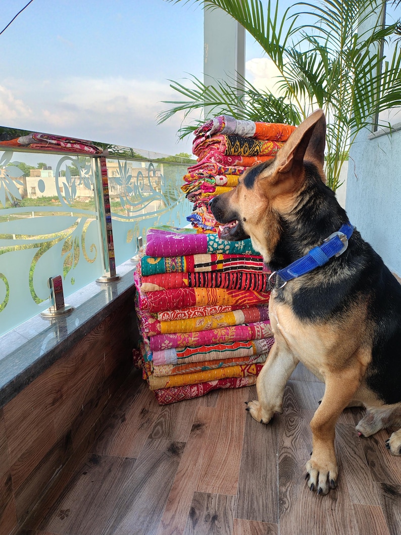 May include: A stack of colorful, patterned quilts in shades of pink, orange, yellow, and purple. A German Shepherd dog with a blue collar sits beside the quilts. The quilts are stacked on a wooden surface.