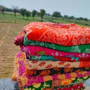 Op de afbeelding: Een stapel kleurrijke, bedrukte quilts. De quilts hebben een verscheidenheid aan kleuren, waaronder oranje, roze, groen en geel. Elke quilt heeft een uniek ontwerp en is netjes op elkaar gestapeld. De quilts zijn gemaakt van een zacht materiaal en lijken handgemaakt.