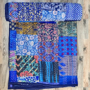 Op de afbeelding: Een patchworkquilt met een verscheidenheid aan blauwe patronen, waaronder bloemen, geometrische en abstracte ontwerpen. De quilt is gemaakt van verschillende stofvierkanten die aan elkaar zijn gestikt, met een donkerblauwe rand. De quilt is bovenaan opgerold.