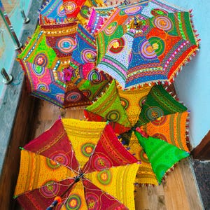 May include: A collection of colorful, decorative umbrellas with intricate patterns. The umbrellas feature a variety of bright colors, including red, yellow, green, and blue, with detailed designs and fringe trim. The umbrellas are open and arranged on a wooden surface.