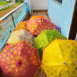 May include: A collection of colorful, open umbrellas with intricate floral patterns. The umbrellas are in shades of yellow, pink, green, and orange, with decorative trim. The scene is set outdoors on a wooden surface.