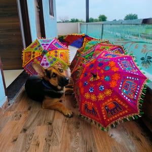 May include: A collection of colorful, decorative umbrellas with intricate embroidered patterns and tassels. The umbrellas feature vibrant hues of pink, yellow, blue, and green. A German Shepherd dog is lying on the wooden floor near the umbrellas.
