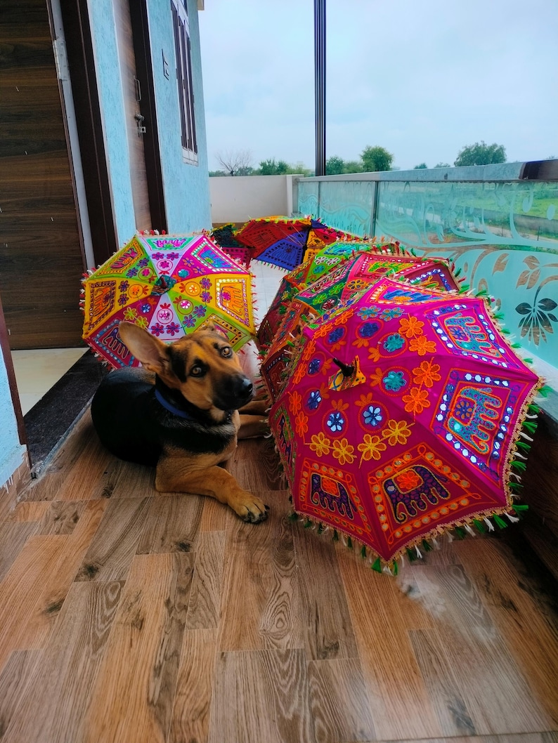 May include: A collection of colorful, decorative umbrellas with intricate embroidered patterns and tassels. The umbrellas feature vibrant hues of pink, yellow, blue, and green. A German Shepherd dog is lying on the wooden floor near the umbrellas.