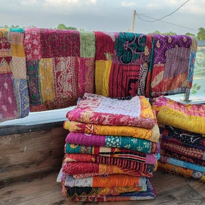 May include: A collection of colorful, patchwork quilts in various sizes. The quilts are displayed folded in stacks and draped over a railing. The quilts feature a variety of patterns and colors, including red, yellow, pink, and blue.