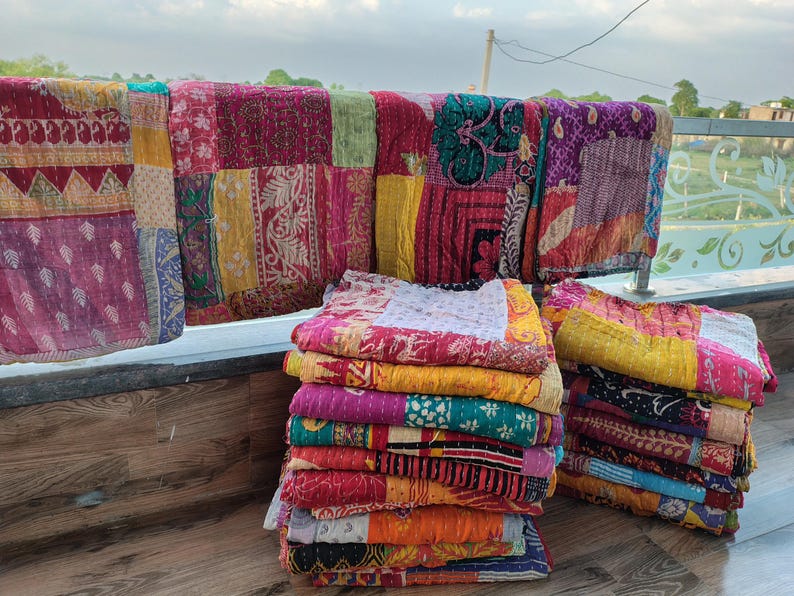 May include: A collection of colorful, patchwork quilts in various sizes. The quilts are displayed folded in stacks and draped over a railing. The quilts feature a variety of patterns and colors, including red, yellow, pink, and blue.