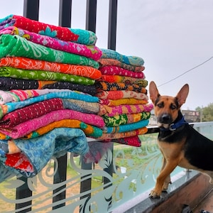 Op de afbeelding: Een stapel kleurrijke, bedrukte quilts in verschillende kleuren, waaronder groen, roze en oranje. Een hond met zwart en bruin haar en een blauwe halsband staat naast de quilts.