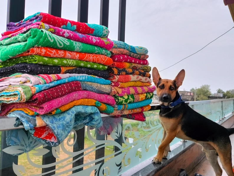 Op de afbeelding: Een stapel kleurrijke, bedrukte quilts in verschillende kleuren, waaronder groen, roze en oranje. Een hond met zwart en bruin haar en een blauwe halsband staat naast de quilts.