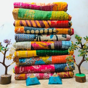 May include: A stack of colorful, patchwork quilts in various patterns and hues, including pink, green, and orange. The quilts are neatly folded and stacked. Decorative miniature trees and blue pyramid-shaped objects are in the foreground.