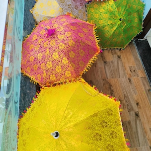 May include: A collection of decorative umbrellas in various colors, including pink, yellow, and green. Each umbrella features a floral pattern and decorative trim. The umbrellas are arranged on a wooden surface.