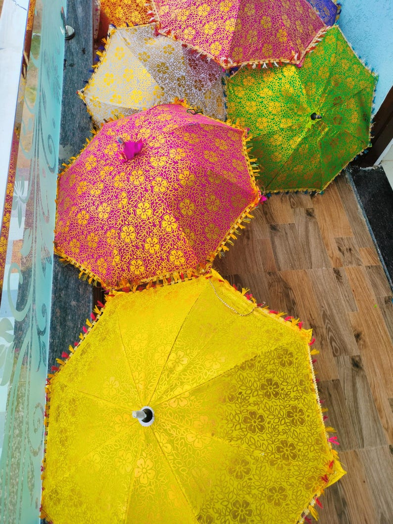 May include: A collection of decorative umbrellas in various colors, including pink, yellow, and green. Each umbrella features a floral pattern and decorative trim. The umbrellas are arranged on a wooden surface.