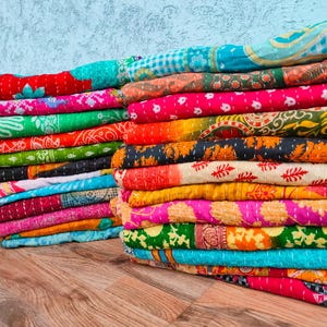 Op de afbeelding: Stapels kleurrijke, bedrukte kantha-quilts. De quilts hebben een verscheidenheid aan levendige kleuren, waaronder roze, groen, oranje en blauw, met ingewikkelde stiksels en bloemmotieven. De quilts zijn opgevouwen en op een houten oppervlak gestapeld.