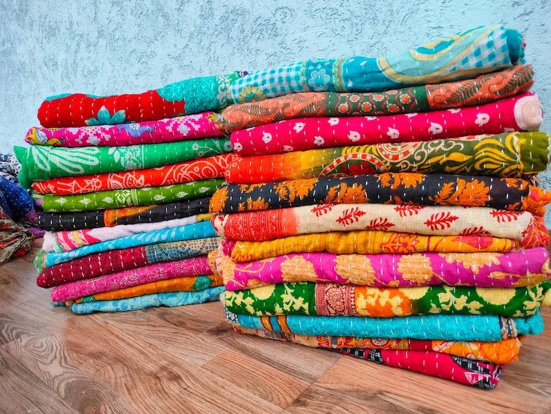 Op de afbeelding: Stapels kleurrijke, bedrukte kantha-quilts. De quilts hebben een verscheidenheid aan levendige kleuren, waaronder roze, groen, oranje en blauw, met ingewikkelde stiksels en bloemmotieven. De quilts zijn opgevouwen en op een houten oppervlak gestapeld.