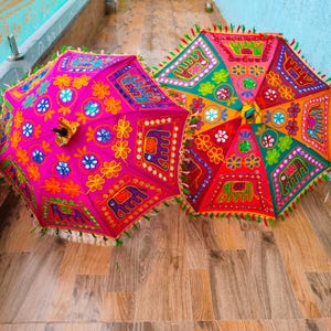 May include: Two colorful, decorative umbrellas with intricate embroidered patterns. One is bright pink, the other is red and orange. Both have detailed designs with floral and elephant motifs, and fringed edges.