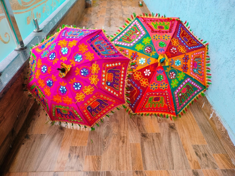 May include: Two colorful, decorative umbrellas with intricate embroidered patterns. One is bright pink, the other is red and orange. Both have detailed designs with floral and elephant motifs, and fringed edges.