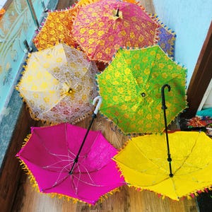 May include: A collection of colorful, decorative umbrellas with intricate floral patterns. The umbrellas are in various shades, including pink, yellow, green, and orange, and are trimmed with tassels. The handles are black or white.