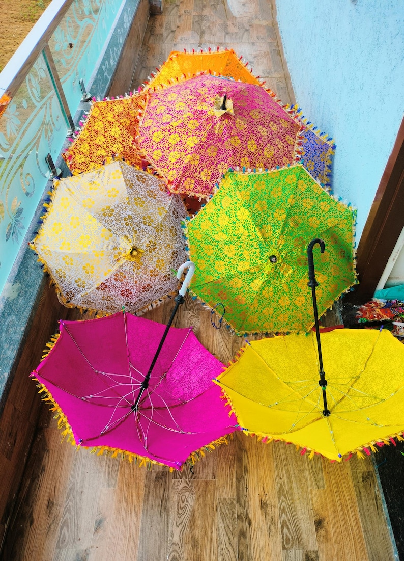 May include: A collection of colorful, decorative umbrellas with intricate floral patterns. The umbrellas are in various shades, including pink, yellow, green, and orange, and are trimmed with tassels. The handles are black or white.
