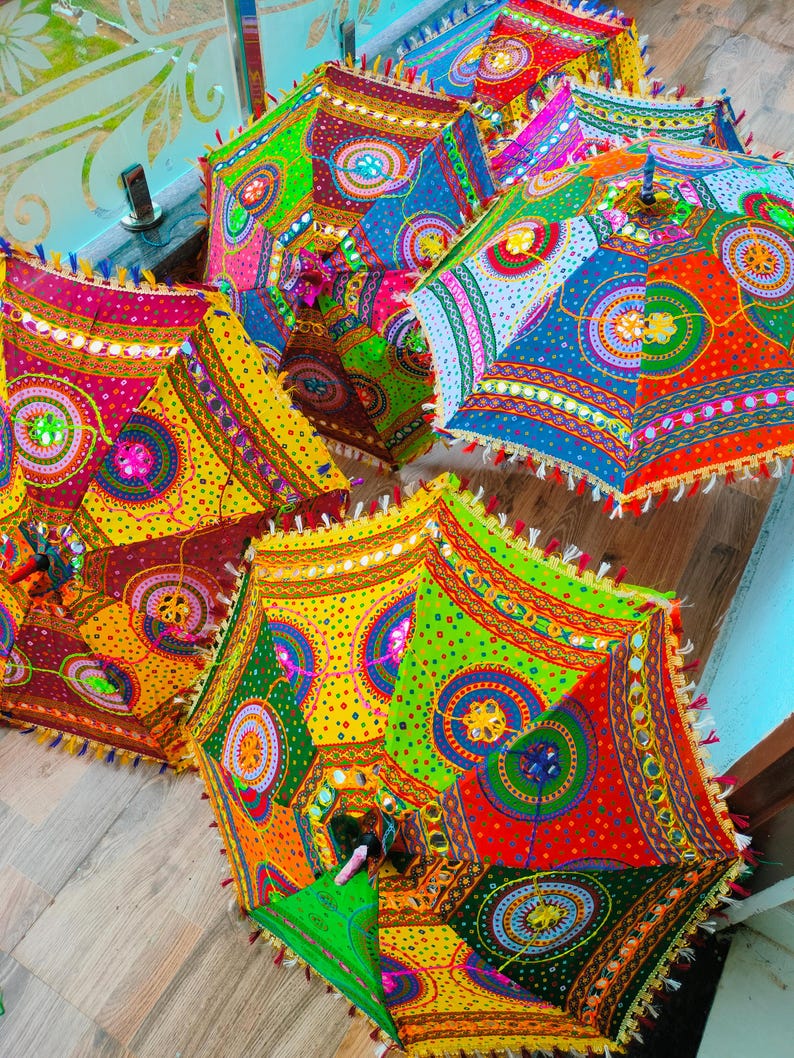 May include: A collection of colorful, patterned umbrellas. Each umbrella features a vibrant patchwork design with red, yellow, green, and blue hues. The edges are trimmed with tassels and small mirrors, creating a festive and decorative look.