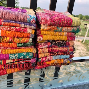 Op de afbeelding: Stapels kleurrijke patchworkquilts in roze, oranje, geel en paars. De quilts zijn gemaakt van verschillende stoffen en patronen, met zichtbare stiksels. De foto is buiten genomen, met een veld op de achtergrond.