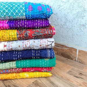 May include: A stack of colorful, quilted blankets. The blankets feature a variety of patterns and colors, including blue, purple, yellow, red, white, green, and pink. Each blanket is stitched with white thread, creating a textured appearance. The blankets are stacked on a wooden surface.