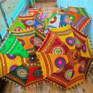 May include: A collection of colorful, decorative umbrellas with intricate patterns and embellishments. The umbrellas feature a vibrant mix of colors, including red, yellow, green, and blue, with detailed mirror work and tassel trim. The umbrellas are open and displayed on a wooden surface.