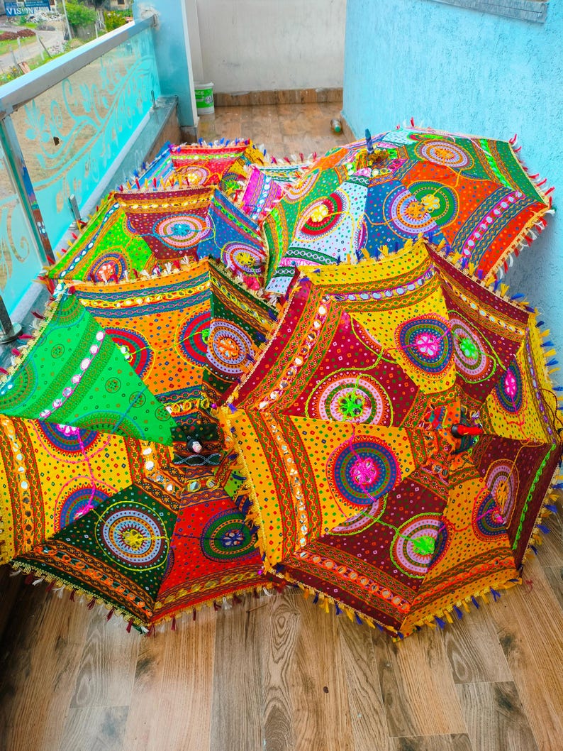 May include: A collection of colorful, decorative umbrellas with intricate patterns and embellishments. The umbrellas feature a vibrant mix of colors, including red, yellow, green, and blue, with detailed mirror work and tassel trim. The umbrellas are open and displayed on a wooden surface.