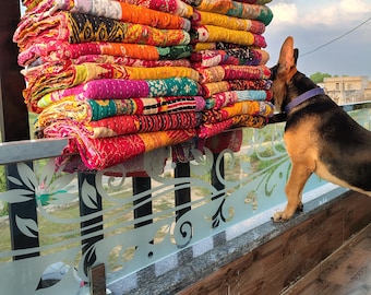 May include: A pile of vibrant, patterned quilts in red, orange, yellow, green, and purple. The quilts are stacked on a decorative railing, with a dog observing them.