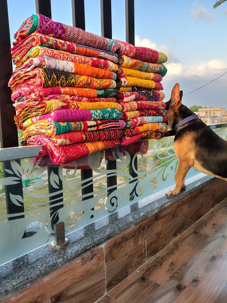 May include: A large stack of colorful, patterned quilts in shades of red, orange, yellow, green, and purple. The quilts are stacked on a decorative railing, with a dog looking at the quilts.