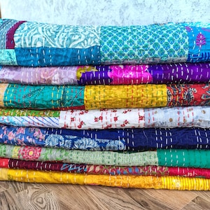 May include: A stack of colorful, patchwork quilts. The quilts feature a variety of patterns and colors, including blue, purple, yellow, and red. Each quilt is made of different fabric pieces sewn together with visible stitching.