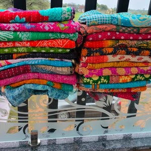 Op de afbeelding: Twee stapels kleurrijke, bedrukte quilts. De quilts zijn opgevouwen en opgestapeld en tonen een verscheidenheid aan levendige kleuren, waaronder rood, groen, blauw en oranje. De quilts lijken gemaakt te zijn van een zachte, gewatteerde stof.