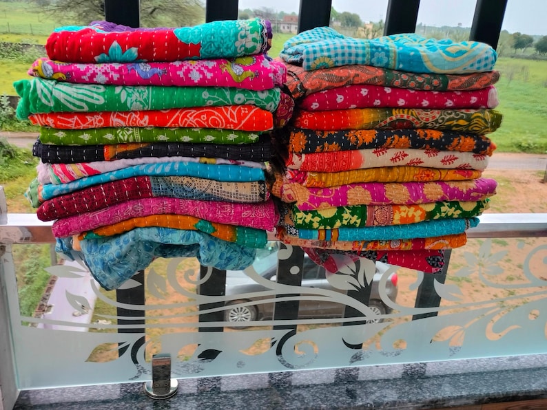 Op de afbeelding: Twee stapels kleurrijke, bedrukte quilts. De quilts zijn opgevouwen en opgestapeld en tonen een verscheidenheid aan levendige kleuren, waaronder rood, groen, blauw en oranje. De quilts lijken gemaakt te zijn van een zachte, gewatteerde stof.