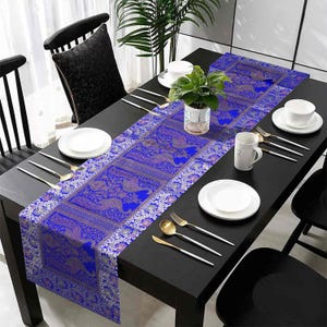 Peut inclure: Un chemin de table bleu et argent avec un motif de paon. Le chemin de table est posé sur une table noire avec des assiettes blanches et des couverts.