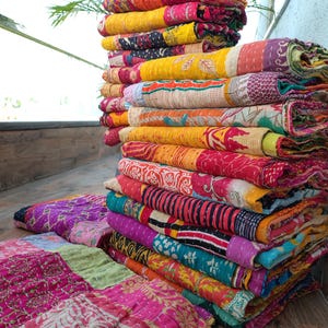 May include: A stack of colorful, patterned quilts. The quilts feature a variety of colors including pink, yellow, orange, and purple. The quilts are folded and stacked on top of each other, with some spread out on the floor.