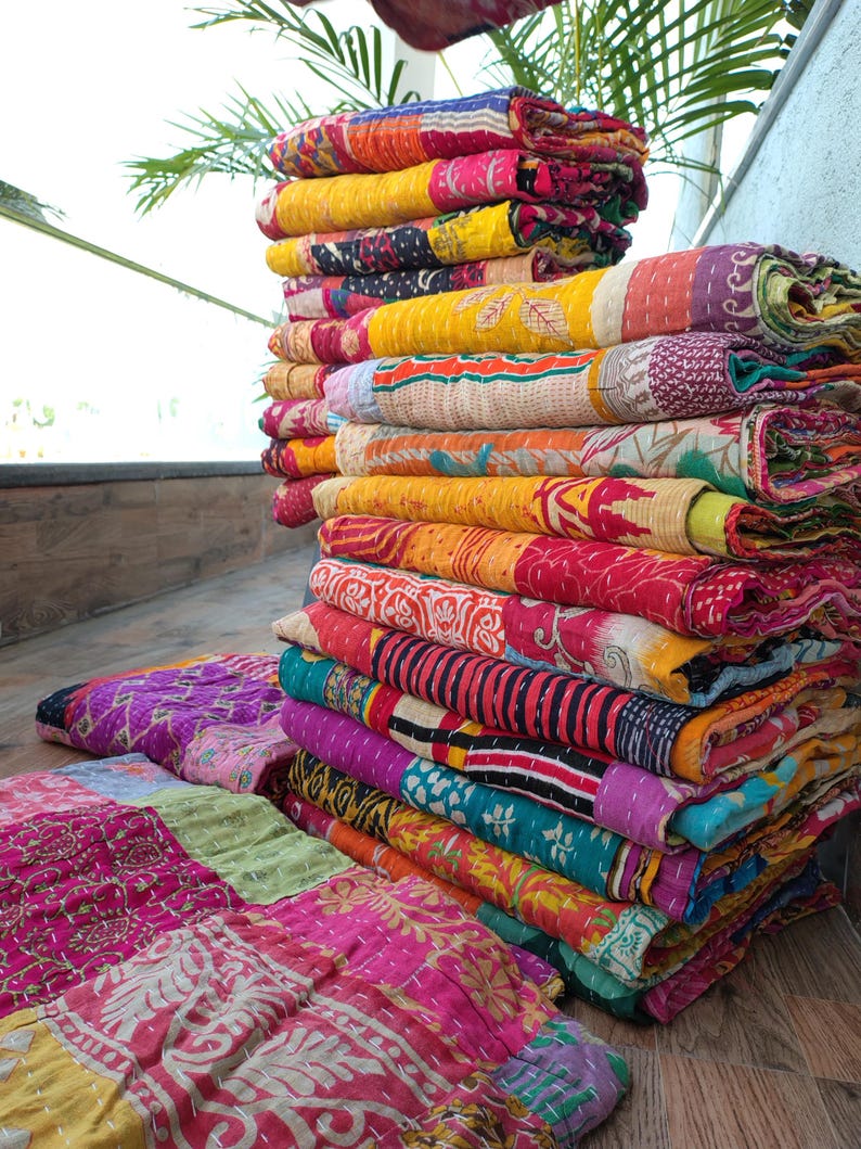 May include: A stack of colorful, patterned quilts. The quilts feature a variety of colors including pink, yellow, orange, and purple. The quilts are folded and stacked on top of each other, with some spread out on the floor.