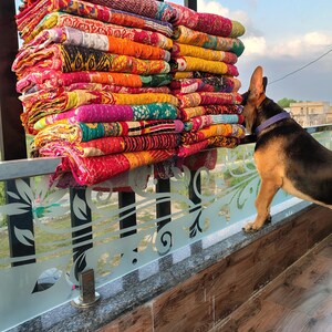 May include: A large stack of colorful, patterned quilts in shades of red, orange, yellow, green, and purple. The quilts are stacked on a decorative railing, with a dog looking at the quilts.