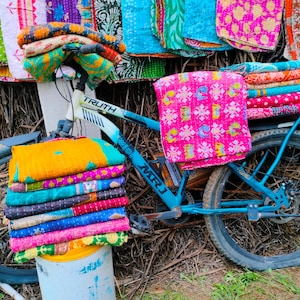Op de afbeelding: Een blauwe fiets met stapels kleurrijke, bedrukte textiel. De textiel is in verschillende kleuren, waaronder roze, geel, groen en blauw. Op de fiets staat het woord "TRUTH".