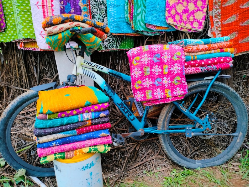 Op de afbeelding: Een blauwe fiets met stapels kleurrijke, bedrukte textiel. De textiel is in verschillende kleuren, waaronder roze, geel, groen en blauw. Op de fiets staat het woord "TRUTH".